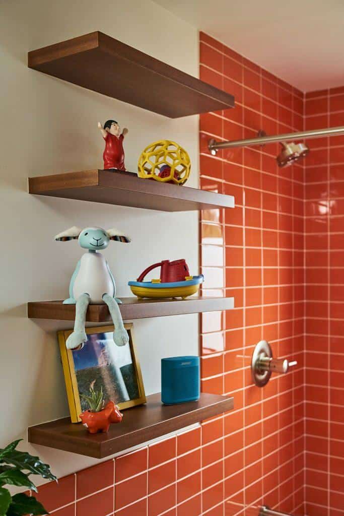 Three wooden shelves on a bathroom wall displaying various items including toys, a framed photo, a small speaker, and decorative objects. The background features orange tiled walls and a showerhead. Three wooden shelves on a bathroom wall displaying various items including toys, a framed photo, a small speaker, and decorative objects. The background features orange tiled walls and a showerhead.