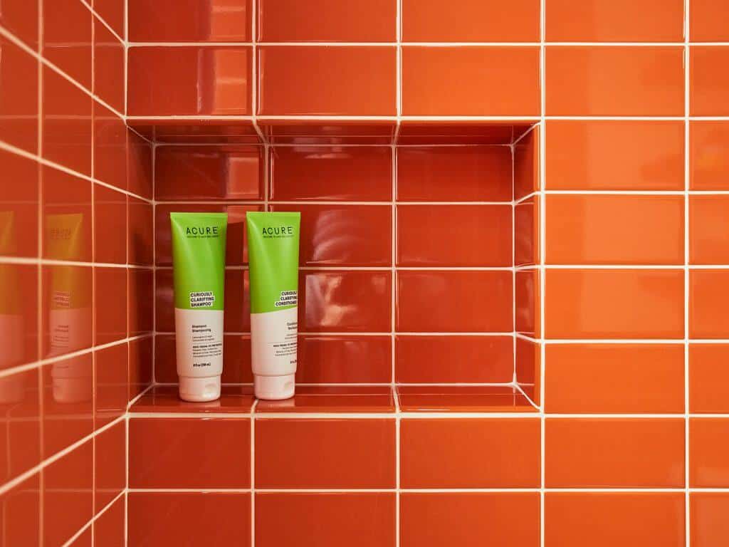 Two green-labeled tubes of Acure shampoo and conditioner are placed on a small built-in shelf in a shower with orange tile walls. Two green-labeled tubes of Acure shampoo and conditioner are placed on a small built-in shelf in a shower with orange tile walls.