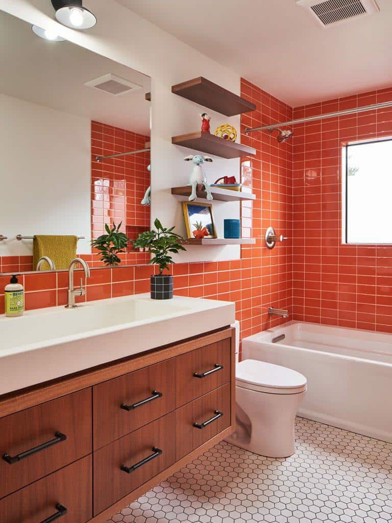 A bathroom with orange tiled walls, a wooden vanity with a white countertop, a modern faucet, a bathtub, a toilet, floating shelves, plants, and a window. A bathroom with orange tiled walls, a wooden vanity with a white countertop, a modern faucet, a bathtub, a toilet, floating shelves, plants, and a window.
