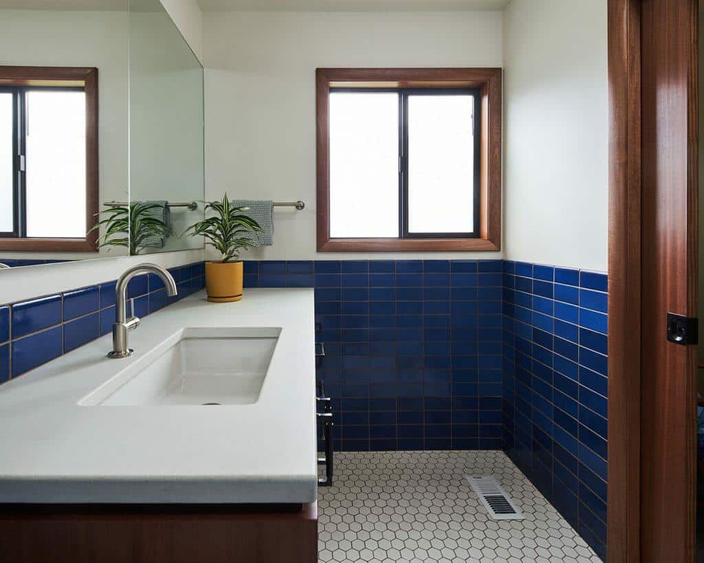 A bathroom with white countertops, a rectangular sink, blue tiled walls, a large mirror, and a window. A potted plant is placed on the counter. The floor features white hexagonal tiles. A bathroom with white countertops, a rectangular sink, blue tiled walls, a large mirror, and a window. A potted plant is placed on the counter. The floor features white hexagonal tiles.