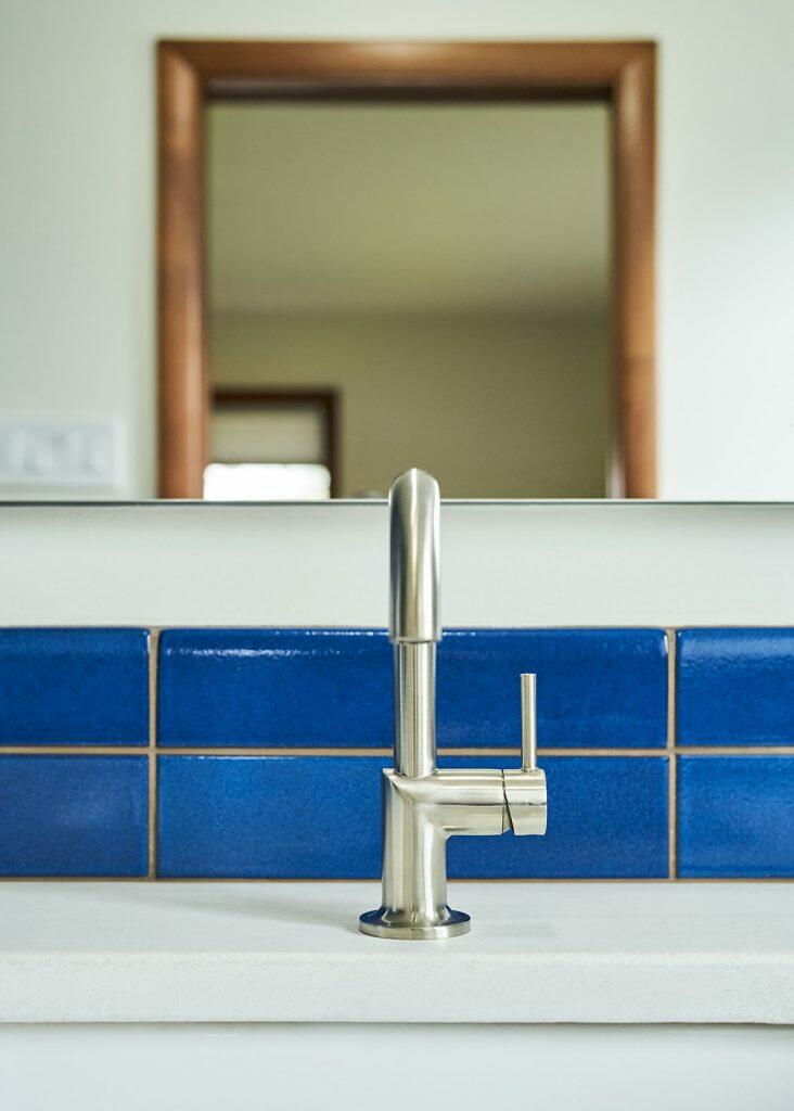 A modern silver faucet on a white countertop, positioned in front of a blue tiled backsplash. A wooden-framed mirror is visible in the background. A modern silver faucet on a white countertop, positioned in front of a blue tiled backsplash. A wooden-framed mirror is visible in the background.