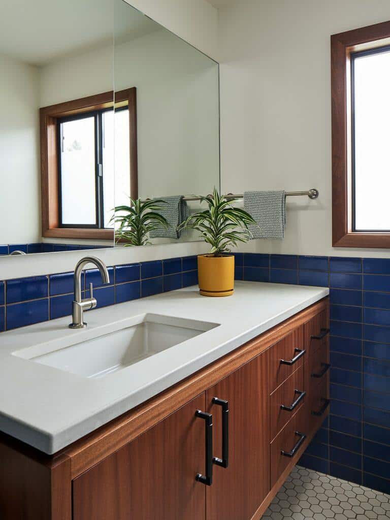 A bathroom features a white sink, wooden vanity with drawers, blue tiled backsplash, and hexagon floor tiles. A small potted plant, a towel on a rail, and two windows with wooden frames are present. A bathroom features a white sink, wooden vanity with drawers, blue tiled backsplash, and hexagon floor tiles. A small potted plant, a towel on a rail, and two windows with wooden frames are present.