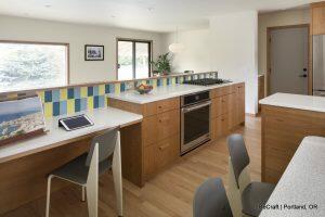 A modern kitchen with light wood cabinets, a gas stove, a tiled backsplash in blue and green, and a white countertop. There is a built-in workspace with a tablet and a chair, and two additional chairs at the counter.