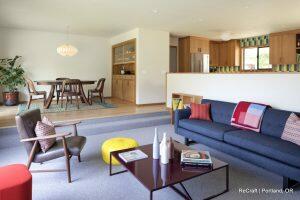 A contemporary living room with a blue sofa, colorful cushions, a modern coffee table, and a lounge chair. The room flows into a dining area with a wooden table and chairs. There's a red ottoman and a yellow pouf.