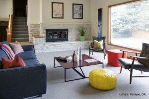 A modern living room with a blue sofa, red table, yellow pouf, black and wood chairs, and decorative items. A large window and stairs are visible. The space features light-colored walls and a stone fireplace.