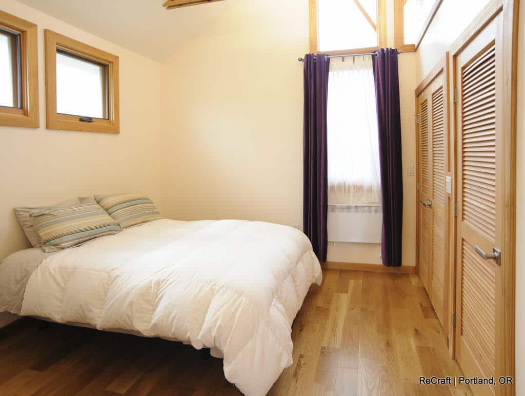 Small bedroom with a cozy bed, wooden flooring, two small windows on the left wall, a large window with curtains on the back wall, and two closed wooden doors on the right. Small bedroom with a cozy bed, wooden flooring, two small windows on the left wall, a large window with curtains on the back wall, and two closed wooden doors on the right.