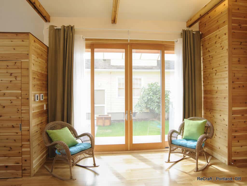 A sunlit room features two wicker chairs with green and blue cushions, wooden walls, and large glass doors opening to an outdoor view. A sunlit room features two wicker chairs with green and blue cushions, wooden walls, and large glass doors opening to an outdoor view.