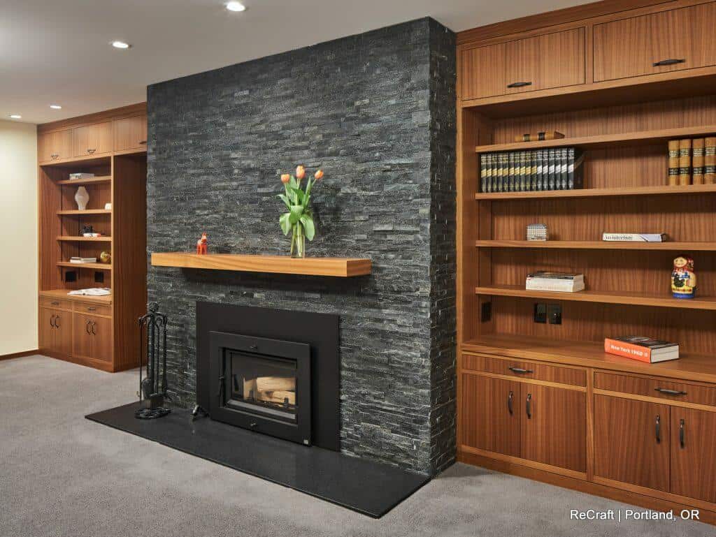 A modern living room features a black stone fireplace centered between wooden shelves filled with books and decorations. A mantel with a vase of flowers and a small figure adorns the fireplace. A modern living room features a black stone fireplace centered between wooden shelves filled with books and decorations. A mantel with a vase of flowers and a small figure adorns the fireplace.