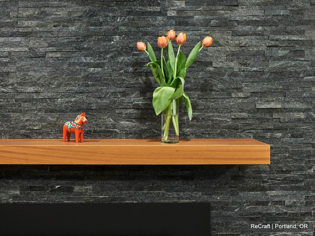 A wooden shelf with an orange horse figurine and a glass vase containing five orange tulips, set against a dark textured stone wall. A wooden shelf with an orange horse figurine and a glass vase containing five orange tulips, set against a dark textured stone wall.