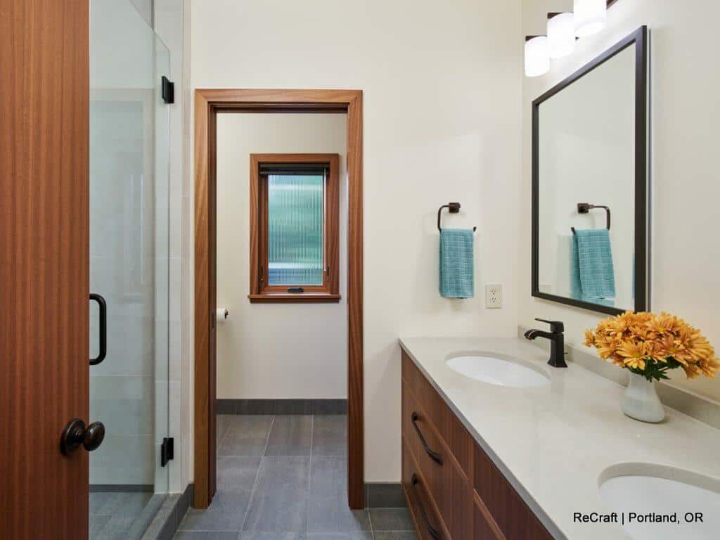 A modern bathroom featuring a double-sink vanity, a glass-enclosed shower, and a small window. A towel hangs by the mirror, and a vase of yellow flowers is placed on the countertop. A modern bathroom featuring a double-sink vanity, a glass-enclosed shower, and a small window. A towel hangs by the mirror, and a vase of yellow flowers is placed on the countertop.