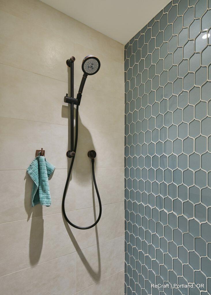 A modern shower with hexagonal teal tiles on one wall, beige tiles on the other, a black handheld showerhead, and a blue towel hanging on a hook. A modern shower with hexagonal teal tiles on one wall, beige tiles on the other, a black handheld showerhead, and a blue towel hanging on a hook.