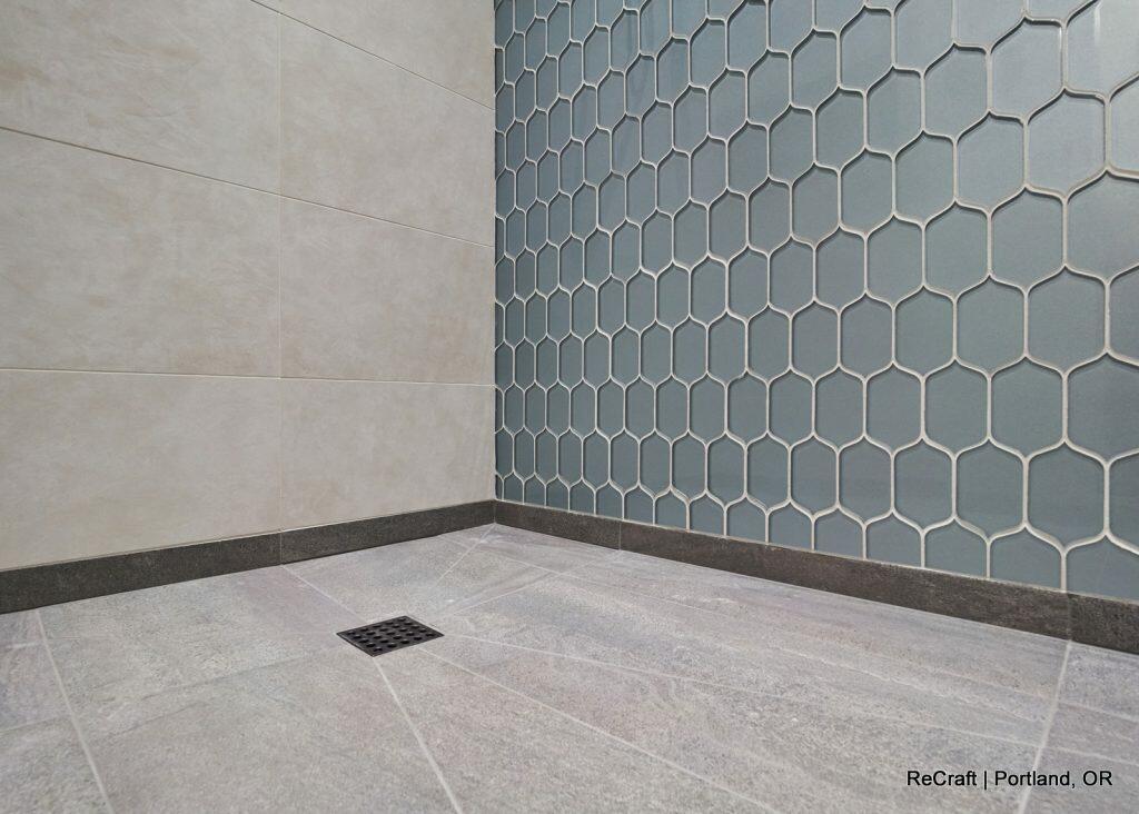 A tiled shower corner with large beige tiles on the left wall, hexagonal blue tiles on the right wall and gray floor tiles with a small square drain. Caption reads "ReCraft | Portland, OR". A tiled shower corner with large beige tiles on the left wall, hexagonal blue tiles on the right wall and gray floor tiles with a small square drain. Caption reads "ReCraft | Portland, OR".