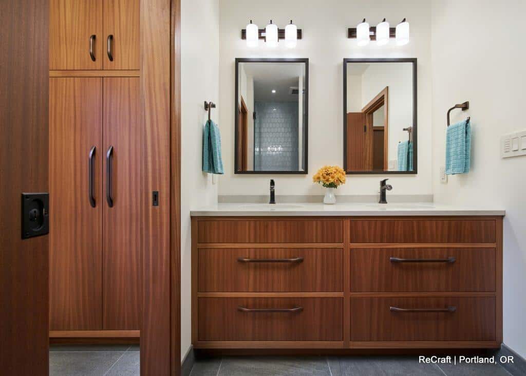 A modern bathroom features a dual-sink vanity with wooden cabinets, two mirrors, and towel racks with blue towels. Overhead lights illuminate the space. A tall cabinet is adjacent on the left. A modern bathroom features a dual-sink vanity with wooden cabinets, two mirrors, and towel racks with blue towels. Overhead lights illuminate the space. A tall cabinet is adjacent on the left.