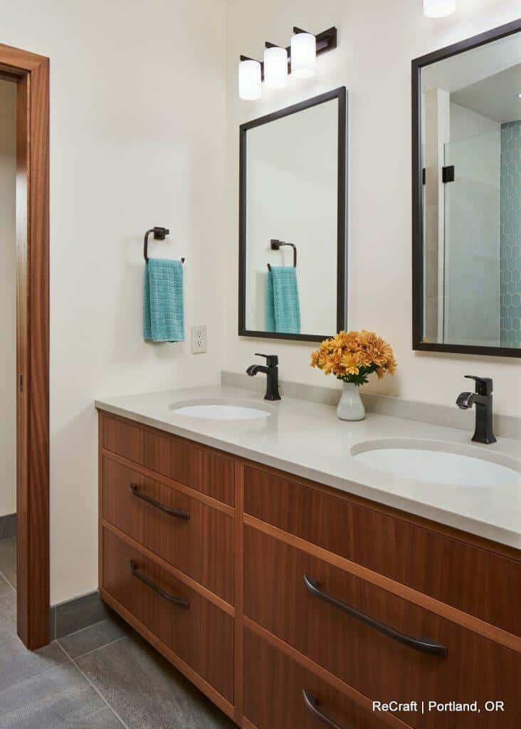 A modern bathroom with double sinks, wooden cabinetry, two wall mirrors, blue hand towels, and a vase of yellow flowers on the countertop. A modern bathroom with double sinks, wooden cabinetry, two wall mirrors, blue hand towels, and a vase of yellow flowers on the countertop.