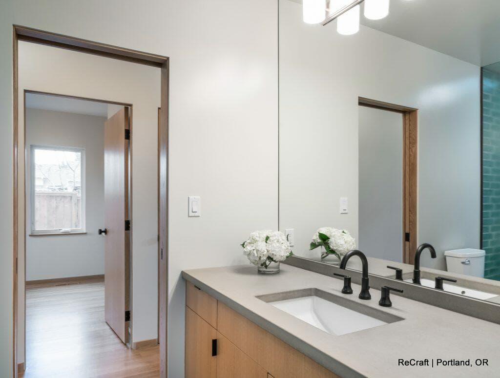 A modern bathroom with a large mirror, dual sinks, black fixtures, and light wood cabinetry. A bouquet of white flowers is on the counter. A view into adjacent room is visible through an open door. A modern bathroom with a large mirror, dual sinks, black fixtures, and light wood cabinetry. A bouquet of white flowers is on the counter. A view into adjacent room is visible through an open door.