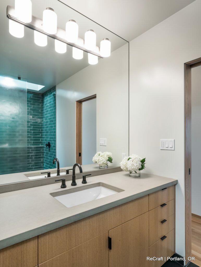 Modern bathroom with a wooden vanity, rectangular sink, black faucet, illuminated mirror, and a glass-enclosed shower with green tiles. Flowers in a vase are on the counter. Modern bathroom with a wooden vanity, rectangular sink, black faucet, illuminated mirror, and a glass-enclosed shower with green tiles. Flowers in a vase are on the counter.