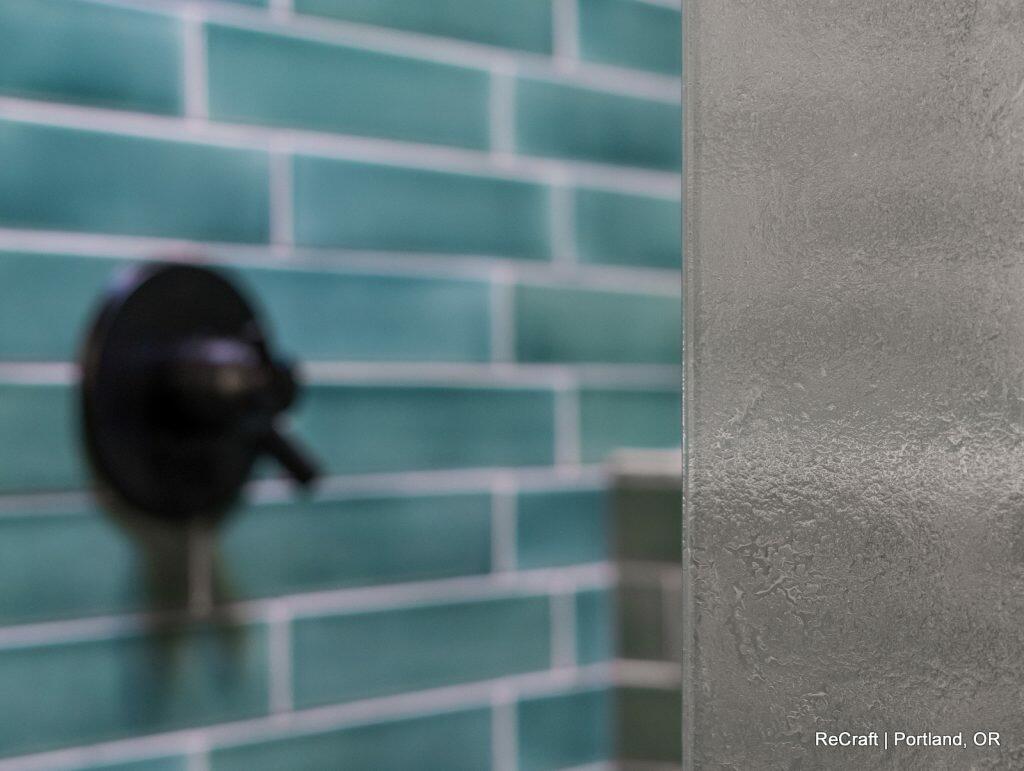 Close-up of a shower area with a frosted glass door, blue-green subway tiles on the wall, and a black shower handle. Text in the lower right corner: "ReCraft | Portland, OR. Close-up of a shower area with a frosted glass door, blue-green subway tiles on the wall, and a black shower handle. Text in the lower right corner: "ReCraft | Portland, OR.