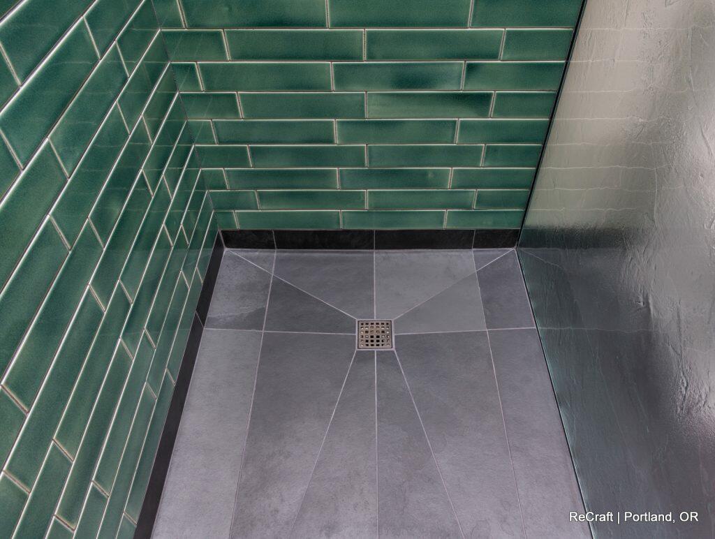 A corner shower with green subway tile walls and grey geometric floor tiles featuring a central square drain. A corner shower with green subway tile walls and grey geometric floor tiles featuring a central square drain.