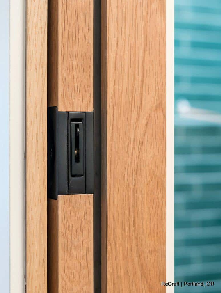Close-up view of a wooden door with a black flush-mounted lock mechanism. Blue-tiled wall is visible in the background. Text at the bottom right corner reads "ReCraft | Portland, OR. Close-up view of a wooden door with a black flush-mounted lock mechanism. Blue-tiled wall is visible in the background. Text at the bottom right corner reads "ReCraft | Portland, OR.