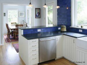 A modern kitchen with blue tile backsplash, stainless steel dishwasher, white cabinetry, and black countertops. In the background, a dining area with a wooden table and chairs is visible.