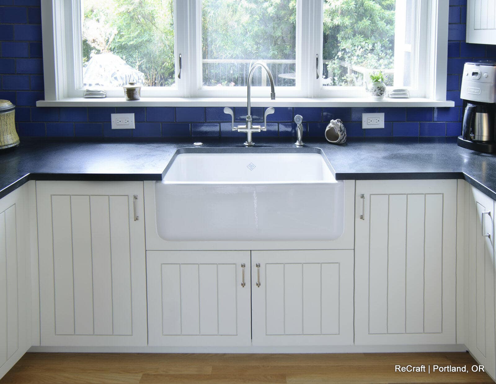 A kitchen with white paneled cabinets, a large farmhouse sink, dark countertops, and a blue tile backsplash, with a window providing natural light. A kitchen with white paneled cabinets, a large farmhouse sink, dark countertops, and a blue tile backsplash, with a window providing natural light.