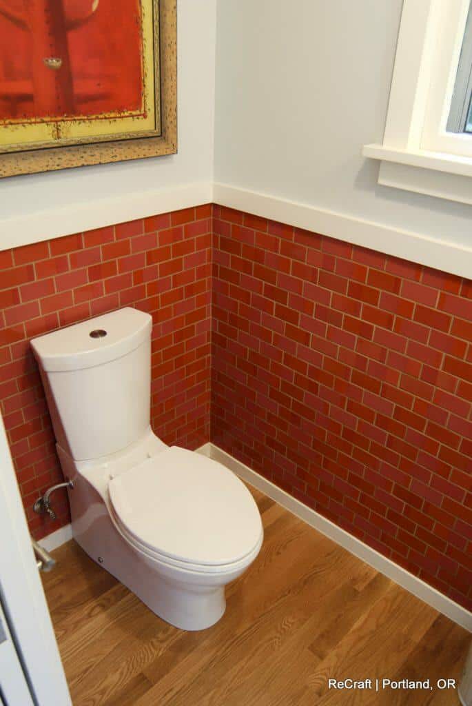 A white toilet situated in a corner with red brick-patterned tile walls and hardwood flooring. A framed picture hangs above the toilet. A white toilet situated in a corner with red brick-patterned tile walls and hardwood flooring. A framed picture hangs above the toilet.