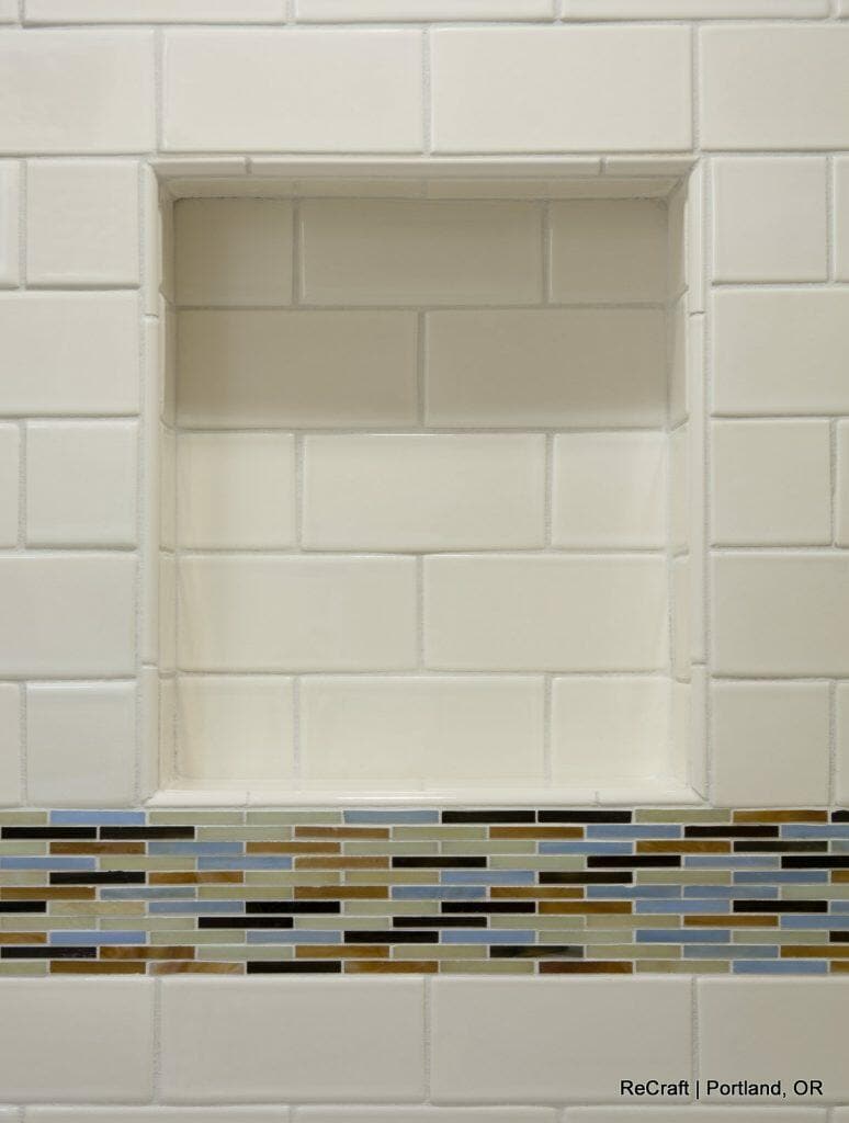 A tiled bathroom niche with a white subway tile background and a horizontal accent strip of small multicolored tiles at the base. A tiled bathroom niche with a white subway tile background and a horizontal accent strip of small multicolored tiles at the base.