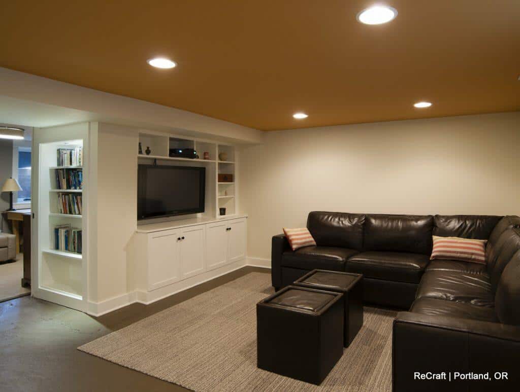 A modern basement living room with a black leather sectional sofa, built-in white shelving with a TV, a beige area rug, and recessed lighting. Adjacent is a small workspace with a bookshelf. A modern basement living room with a black leather sectional sofa, built-in white shelving with a TV, a beige area rug, and recessed lighting. Adjacent is a small workspace with a bookshelf.
