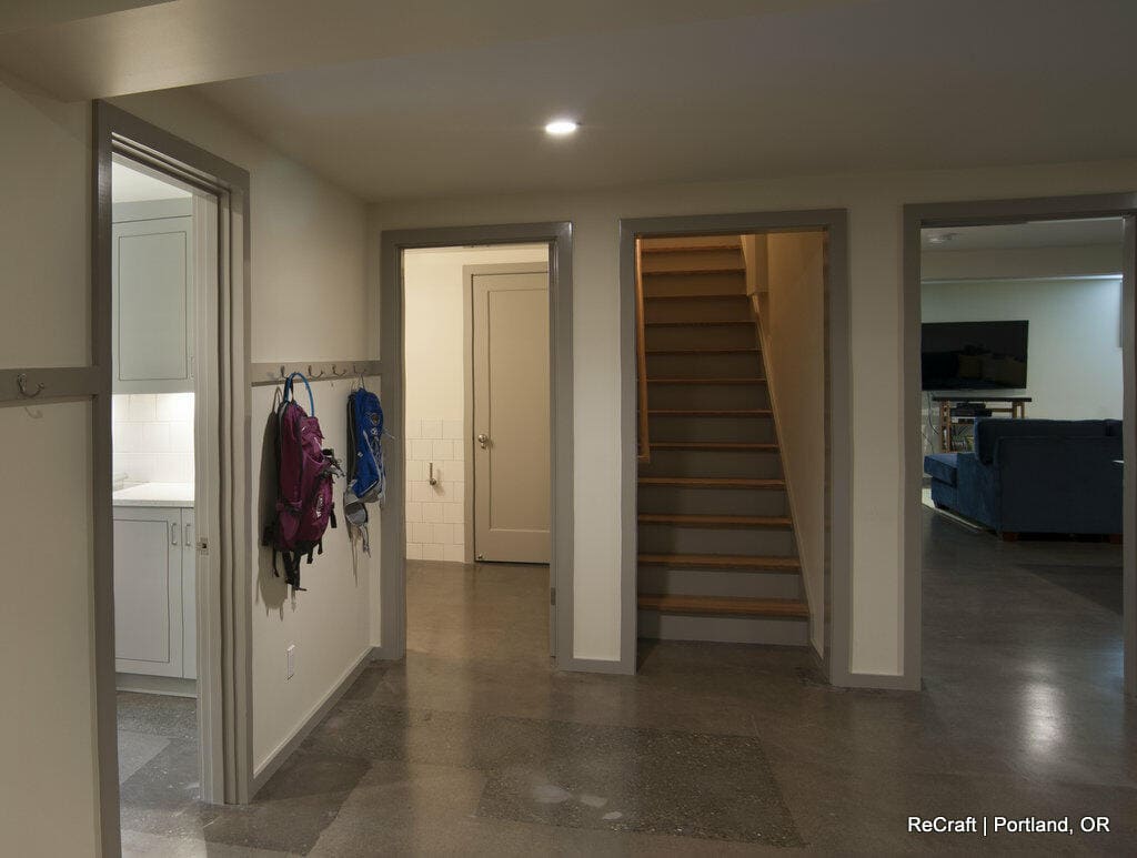 A basement with polished concrete floors and neutral walls includes a laundry area, a staircase, hooks with backpacks, and a living space with a TV and blue sofa. Text: "ReCraft | Portland, OR". A basement with polished concrete floors and neutral walls includes a laundry area, a staircase, hooks with backpacks, and a living space with a TV and blue sofa. Text: "ReCraft | Portland, OR".