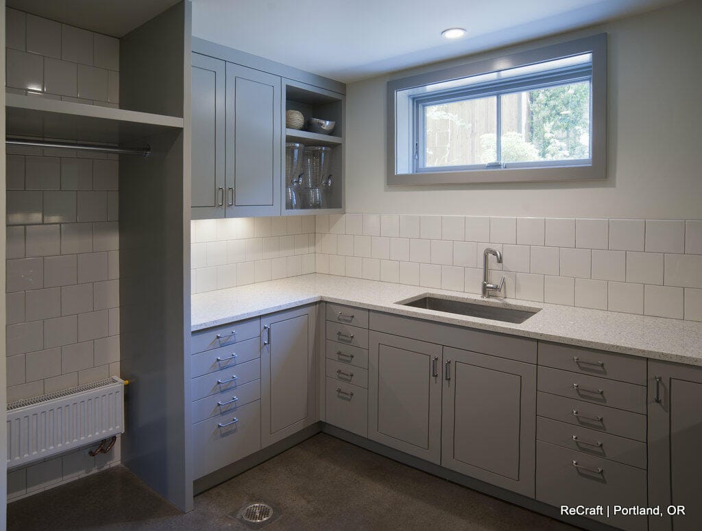 A modern kitchen with gray cabinets, white countertops, a single-basin sink, and a window above. Open shelving displays dishes. A small radiator is beneath an empty shelf on the left. A modern kitchen with gray cabinets, white countertops, a single-basin sink, and a window above. Open shelving displays dishes. A small radiator is beneath an empty shelf on the left.