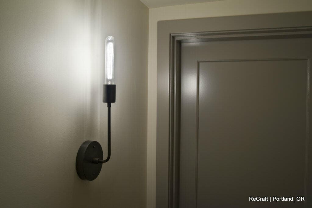 A wall-mounted light fixture with an exposed bulb illuminates a hallway next to a closed door. Text at the bottom right reads, "ReCraft | Portland, OR. A wall-mounted light fixture with an exposed bulb illuminates a hallway next to a closed door. Text at the bottom right reads, "ReCraft | Portland, OR.