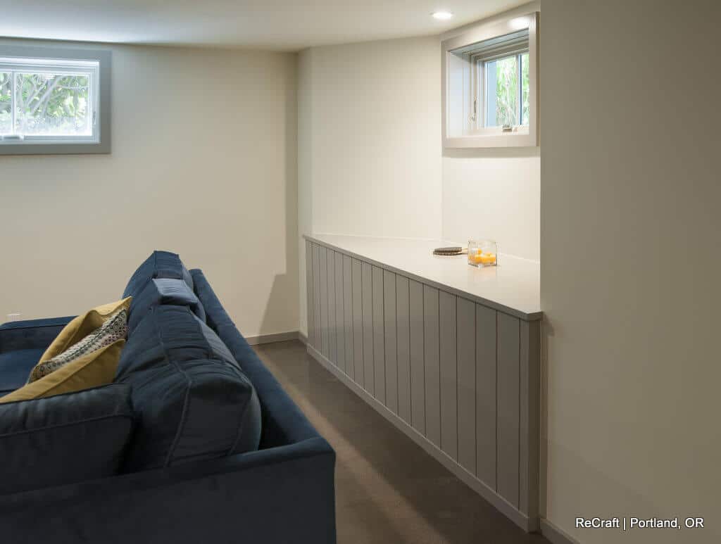 A basement room with a navy-blue sofa, throw pillows, a long white countertop against the wall, and small windows above. The space is clean, modern, and softly lit. A basement room with a navy-blue sofa, throw pillows, a long white countertop against the wall, and small windows above. The space is clean, modern, and softly lit.