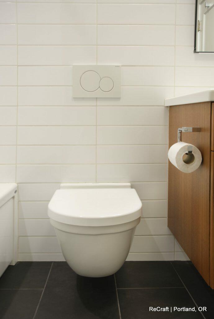 A modern bathroom with white tile walls, a wall-mounted toilet, and a wooden vanity with a toilet paper holder. A modern bathroom with white tile walls, a wall-mounted toilet, and a wooden vanity with a toilet paper holder.