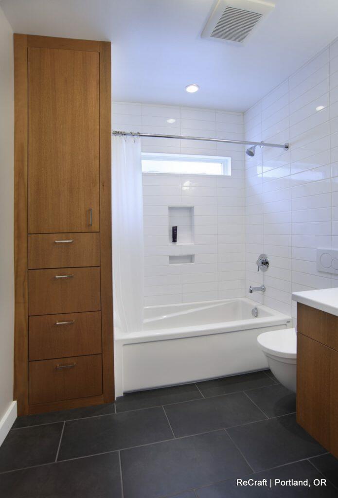 Modern bathroom with white tiled walls, a bathtub with a showerhead, a white curtain, wooden cabinets on the left, and dark floor tiles. Modern bathroom with white tiled walls, a bathtub with a showerhead, a white curtain, wooden cabinets on the left, and dark floor tiles.