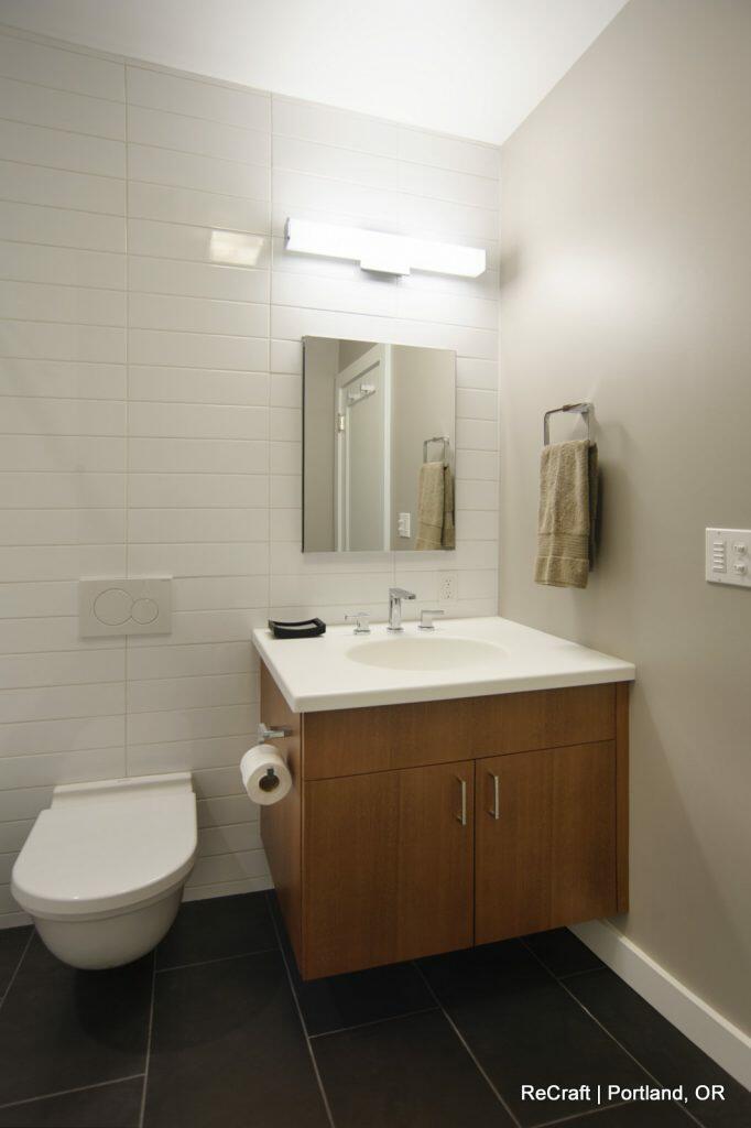 A modern bathroom with a wall-hung toilet, vanity with sink and mirror, white tiles on the wall, a towel ring with a towel, and a dark tile floor. A modern bathroom with a wall-hung toilet, vanity with sink and mirror, white tiles on the wall, a towel ring with a towel, and a dark tile floor.