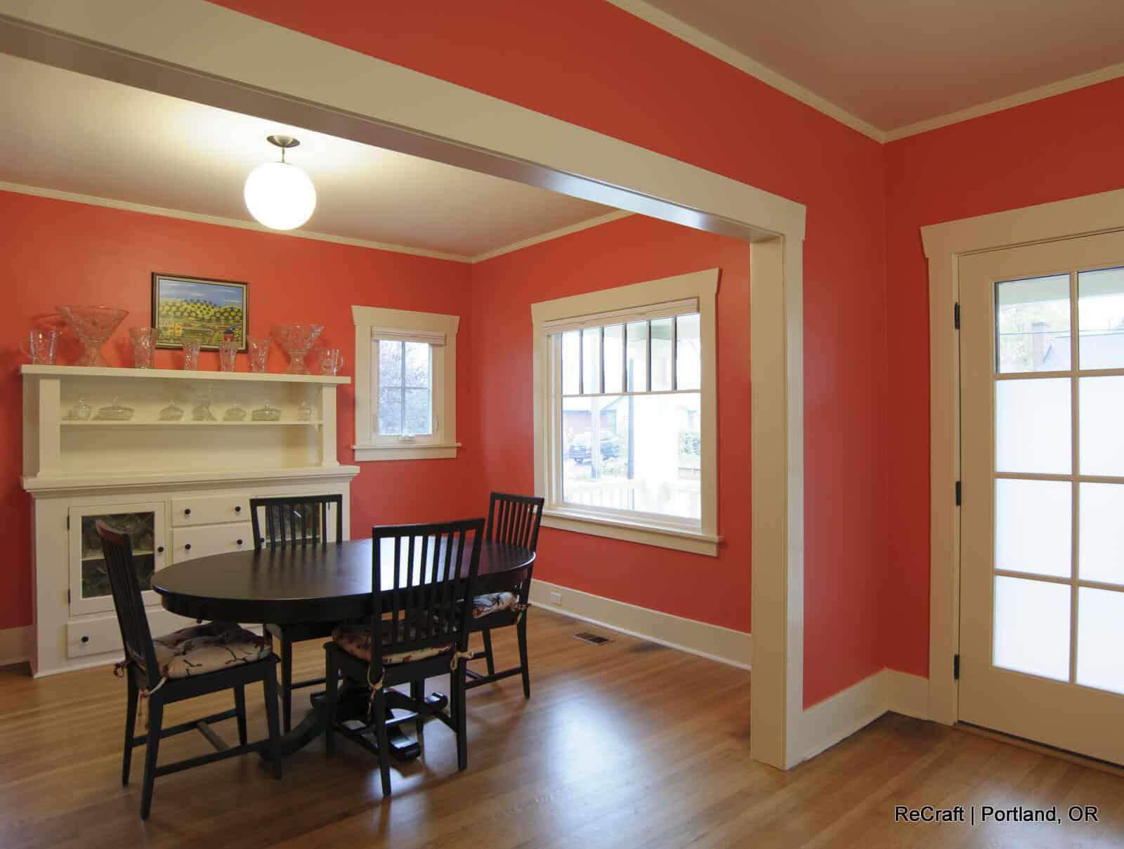 Dining area with a round black table and black chairs, featuring coral walls, a white fireplace, and large windows, showcasing a modern yet inviting home remodeling design by ReCraft Home Remodeling.