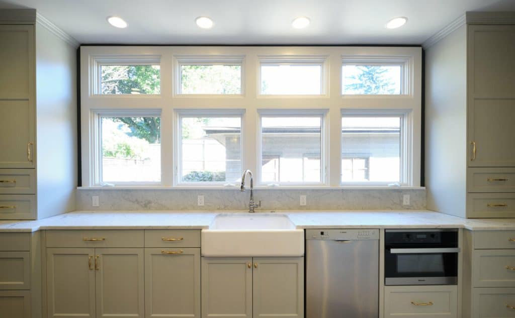Modern kitchen featuring large windows, white farmhouse sink, stainless steel dishwasher, built-in oven, and gray cabinetry with brass handles under ample natural light. Modern kitchen featuring large windows, white farmhouse sink, stainless steel dishwasher, built-in oven, and gray cabinetry with brass handles under ample natural light.