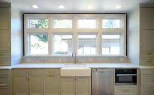 Modern kitchen featuring large windows, white farmhouse sink, stainless steel dishwasher, built-in oven, and gray cabinetry with brass handles under ample natural light.