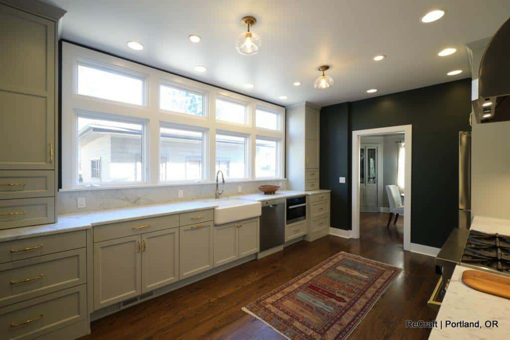 A modern kitchen with light green cabinets, a farmhouse sink, stainless steel appliances, and hardwood floors. Natural light streams through large windows, illuminating a patterned rug on the floor. A modern kitchen with light green cabinets, a farmhouse sink, stainless steel appliances, and hardwood floors. Natural light streams through large windows, illuminating a patterned rug on the floor.