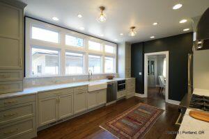 A modern kitchen with light green cabinets, a farmhouse sink, stainless steel appliances, and hardwood floors. Natural light streams through large windows, illuminating a patterned rug on the floor.
