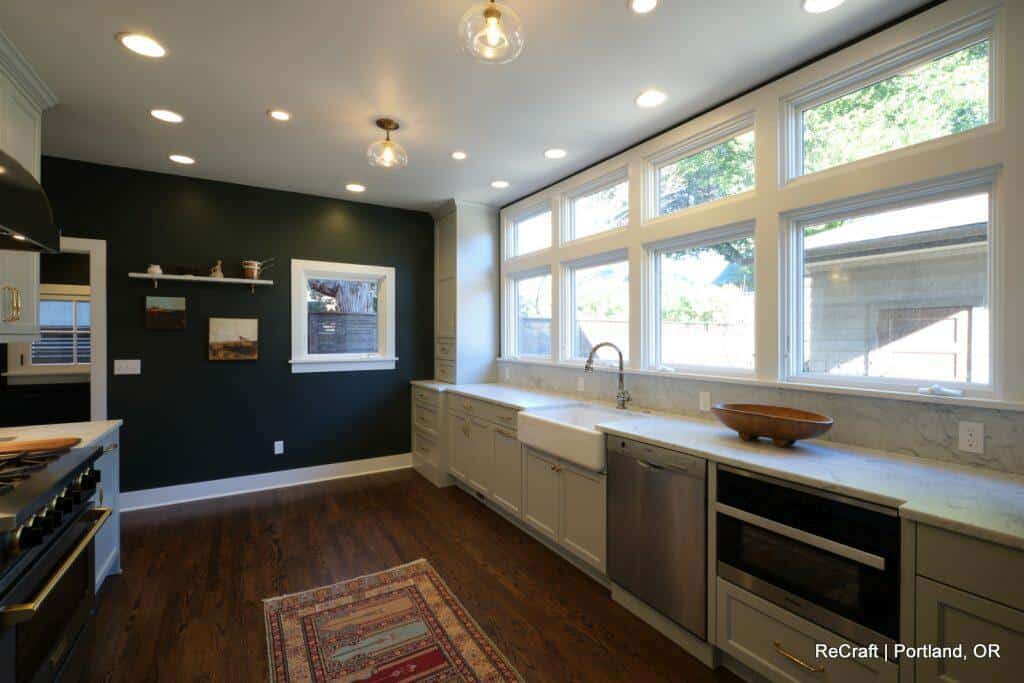 Modern kitchen with dark green accent wall, recessed and pendant lighting, white cabinets, marble countertops, stainless steel appliances, farmhouse sink, and large windows. A rug lies on wooden floor. Modern kitchen with dark green accent wall, recessed and pendant lighting, white cabinets, marble countertops, stainless steel appliances, farmhouse sink, and large windows. A rug lies on wooden floor.