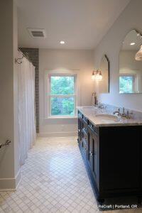 Modern bathroom featuring a large vanity with dual sinks, a window, a shower with a curtain, and geometric tiled flooring. Wall sconce lighting above the mirror and a view of trees outside the window.
