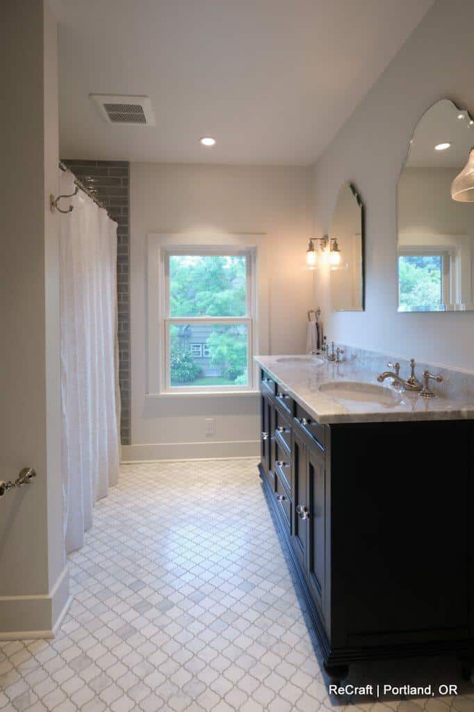 Modern bathroom featuring a large vanity with dual sinks, a window, a shower with a curtain, and geometric tiled flooring. Wall sconce lighting above the mirror and a view of trees outside the window. Modern bathroom featuring a large vanity with dual sinks, a window, a shower with a curtain, and geometric tiled flooring. Wall sconce lighting above the mirror and a view of trees outside the window.