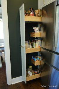 Tall, narrow kitchen pantry with pull-out wooden shelves containing various food items and snacks, next to a stainless steel refrigerator.