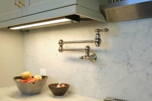 A kitchen with a marble backsplash features a polished metal pot filler faucet mounted on the wall. A bowl of various produce sits on the counter.