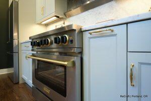 A modern kitchen featuring a stainless steel oven with gold accents, white cabinets, and a marble countertop. There's a black refrigerator in the background.