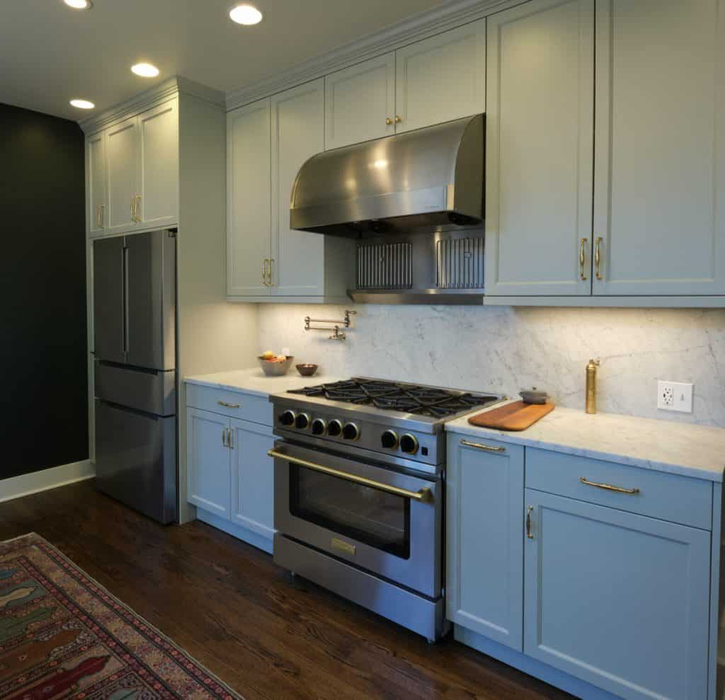 Modern kitchen with stainless steel appliances including a refrigerator and a large oven with ventilation hood, white marble countertops, light blue cabinets, and dark wood flooring. Modern kitchen with stainless steel appliances including a refrigerator and a large oven with ventilation hood, white marble countertops, light blue cabinets, and dark wood flooring.