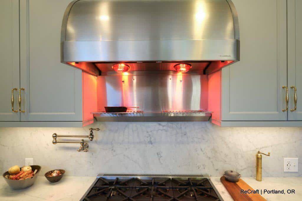 Modern kitchen with a stainless steel range hood, marble backsplash, and gas stove. Various cooking utensils and ingredients are placed on the countertop. Warm lighting illuminates the space. Modern kitchen with a stainless steel range hood, marble backsplash, and gas stove. Various cooking utensils and ingredients are placed on the countertop. Warm lighting illuminates the space.