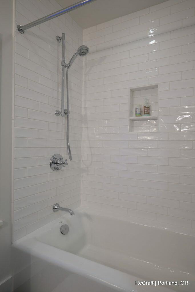 A modern white-tiled bathtub and shower with a chrome showerhead, faucet, and a built-in shelf containing toiletries. A modern white-tiled bathtub and shower with a chrome showerhead, faucet, and a built-in shelf containing toiletries.