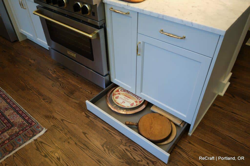 A kitchen with light blue cabinets, stainless steel stove, and a drawer pulled out containing cutting boards and a plate. The floor is wooden, and part of a rug is visible. "ReCraft | Portland, OR" text in the image. A kitchen with light blue cabinets, stainless steel stove, and a drawer pulled out containing cutting boards and a plate. The floor is wooden, and part of a rug is visible. "ReCraft | Portland, OR" text in the image.